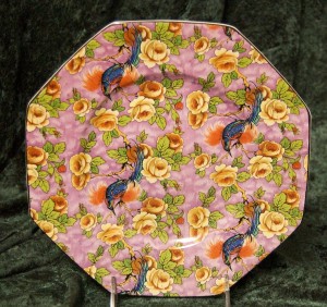 This is a Chintz Plate with Birds and is marked Baker & Co England with the trademark being a standing Lion in a Shield. It is decorated with Exotic Tropical Birds, Yellows Roses over all and has a soft lavender dabbed with a lighter muted tones of very pale lavender over all in the background. It measures 9" and is in a hexagon shape with a black outlined edge.